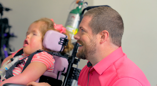 Addie, a child with special needs, sits in her motorized wheelchair as her father Mathew attends to her.