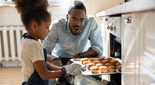 Dad happily supervises his young daughter remove fresh-baked muffins from the oven.