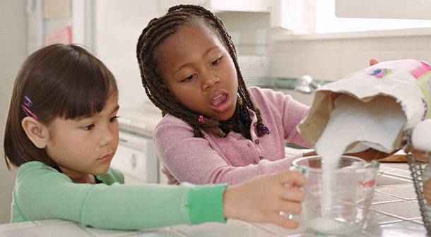 A girl helps her friend measure sugar for a recipe.